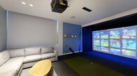 Located at 201 Clearwater Dr in West Palm Beach, FL, this single-floor sports simulator room is a purpose-built entertainment space designed for immersive athletic gaming experiences. The room is anchored by a professional-grade multi-sport simulator, complete with a large projection screen, blue ambient lighting, and a dedicated artificial turf hitting mat — ideal for golf, baseball, and a variety of other simulated sports. A ceiling-mounted projector delivers crisp, large-format visuals, while the darkened simulator bay creates a focused, stadium-like atmosphere. A blue accent wall adorned with mounted sports balls and a baseball bat adds personality and reinforces the sporting theme. The lounge area features a generously sized light grey sectional sofa paired with organic-shaped wooden and yellow coffee tables, offering comfortable viewing for spectators. Recessed ceiling lighting keeps the space well-lit outside the simulator bay. A wood-toned media console with a wall-mounted flat-screen TV and computer setup provides additional control and viewing options. The flooring transitions between plush carpet in the lounge area and dark turf in the simulator zone, clearly delineating function within the space. Clean white walls and a modern, streamlined design complete the polished, contemporary aesthetic.