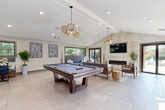 This well-appointed amenity space is thoughtfully designed to offer a variety of social and recreational experiences under one roof. The interior is anchored by soaring vaulted ceilings with exposed beam detailing and large-format porcelain tile flooring that runs seamlessly throughout, creating a clean, cohesive foundation for the space.

The main gathering area centers around a full-size billiards table positioned beneath a pair of sculptural pendant light fixtures with woven rattan shades, adding warmth and artistic flair to the room. Adjacent to the billiards area, a comfortable lounge zone features wood-framed sofas and armchairs upholstered in cool blue-grey tones, arranged around a textured area rug and woven side tables.

A floor-to-ceiling stone-clad fireplace wall serves as a dramatic focal point, with a wall-mounted television above a sleek linear fireplace. Sliding glass doors and expansive dark-framed windows flood the space with natural light and offer views of the surrounding lush greenery and outdoor areas.

A built-in banquette seating nook with navy upholstery and bistro-style tables provides an intimate dining or casual meeting area. Nearby, a kitchenette features herringbone tile backsplash, dark wood cabinetry, and stainless steel appliances. Abstract wall art and potted greenery complete the modern coastal aesthetic throughout.