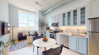 This single-floor unit showcases a thoughtfully designed open-concept layout that seamlessly integrates the kitchen, dining, and living areas. The kitchen is a standout feature, outfitted with tall shaker-style cabinetry in soft white with ribbed glass upper panels, a marble-look tile backsplash with under-cabinet lighting, and a full suite of stainless steel appliances including a range, microwave, dishwasher, and refrigerator. An exposed metallic ductwork runs overhead, lending an industrial character that contrasts elegantly with the clean, bright finishes throughout. Light wood-tone flooring and a ceiling fan enhance the airy feel of the open living and dining space, which is anchored by a large window offering scenic outdoor views and ample natural light. The bedroom features a bold deep-blue accent wall, creating a cozy and stylish retreat complemented by warm ambient lighting. The bathroom is finished in neutral tones with large-format floor tiles, a wall-mounted floating vanity with backlit mirror, and a glass-enclosed walk-in shower, delivering a spa-like atmosphere. Overall, the space balances modern urban design with warm, livable comfort.