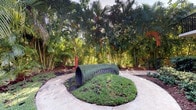 This single-level outdoor space showcases a distinctive grass-covered cylindrical tunnel structure positioned at the center of a concrete pad, creating a playful and naturalistic focal point. The tunnel is wrapped in artificial or natural turf, blending organically with the surrounding ground cover that spills outward from its base. A red vertical post stands adjacent to the structure, likely serving a functional or boundary-marking purpose.

The space is enveloped by a dense, mature tropical landscape featuring towering palm trees, bamboo clusters, and a rich variety of lush greenery that creates a sense of seclusion and natural immersion. The vegetation forms a layered canopy overhead, filtering natural light softly throughout the space. The overall ambiance is tropical and verdant, suggesting a warm climate environment.

The concrete paved area provides a clean, structured surface that contrasts with the organic forms of the surrounding plantings. A metal fence is partially visible along the perimeter, defining the boundary of the space. This outdoor area appears well-suited for recreational or interactive use, combining hardscape and landscape elements thoughtfully within a tropical setting.
