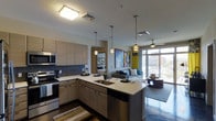 This single-floor unit showcases a thoughtfully designed open-plan layout that seamlessly integrates the kitchen and living areas. The kitchen is appointed with flat-front cabinetry in a warm wood-tone finish, complemented by a mosaic tile backsplash and light quartz countertops. A central island with an undermount sink provides additional prep space and casual dining, illuminated by a series of stylish pendant lights in varying designs. Stainless steel appliances, including a built-in microwave, electric range, and dishwasher, complete the functional cooking space.

The living area flows naturally from the kitchen, anchored by dark hardwood-style flooring that extends throughout. Bold, large-scale artwork adds a vibrant, artistic character to the space. Floor-to-ceiling windows along the far wall flood the interior with natural light while offering urban views and balcony access, framed by yellow accent curtains. A ceiling fan enhances comfort in the living zone. Pendant and flush-mount lighting throughout ensures the space is well-lit, contributing to an overall modern, urban aesthetic.