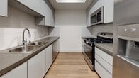 This contemporary apartment showcases a thoughtful blend of form and function. The open-concept living area is anchored by a sleek, minimalist kitchen featuring white cabinetry, stainless steel appliances, and a practical galley layout. Recessed lighting and a tray ceiling add depth to the space.

The living room is furnished with a comfortable grey sofa and a stylish wooden coffee table, complemented by an accent chair in warm tones. Light wood-look flooring flows throughout, creating a cohesive aesthetic. Large windows in the bedroom and living areas flood the space with natural light, while offering views of the surrounding area.

The bedroom is spacious, accommodating a queen-sized bed and featuring a mirrored sliding closet door. An in-unit laundry closet with a stacked washer and dryer adds convenience to daily living. The bathroom continues the modern theme with a floating vanity, large mirror, and a glass-enclosed shower.

Overall, this apartment combines contemporary design with practical amenities, creating an inviting and efficient living space. The neutral color palette and clean lines contribute to a sense of spaciousness and calm throughout the unit.