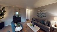 This single-story residence is thoughtfully laid out across several functional spaces, each finished with a consistent neutral color palette of warm grays and taupe tones complemented by teal and blue accents throughout. The living room provides a comfortable gathering space with carpet flooring, a textured ceiling, and a sliding glass door fitted with vertical blinds that opens to an outdoor patio, allowing natural light to filter into the room. Adjacent to the living area, a dedicated dining space accommodates a round table and seating for four beneath a pendant light, positioned directly alongside the kitchen for convenient flow. The galley-style kitchen is efficiently organized with dark cabinetry on both sides, white countertops, track lighting overhead, hardwood-style flooring, and a full suite of white appliances including a range and refrigerator. The primary bedroom features ceiling fan lighting, soft carpet, a walk-in closet, and a window with blinds. Additional bedrooms offer similar finishes with ceiling fans and carpeted floors, suitable for sleeping or as a dedicated workspace. The bathroom area includes a dual-sink vanity with dark cabinetry and white countertops, wood-style flooring, and a separate water closet. Throughout the residence, textured ceilings and a cohesive design create a unified and functional living environment.