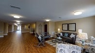 Located in Rolesville, NC, this single-story space offers an open and welcoming layout centered around a spacious living area. The room features warm hardwood flooring that runs throughout, complementing the neutral tan and beige wall tones that create a calm, inviting atmosphere. Flush-mount ceiling lighting provides consistent illumination across the open floor plan.

The living area is thoughtfully arranged with a dark upholstered sofa accented by teal and gray throw pillows, paired with a rustic wooden bench and a classic rocking chair, offering multiple seating options. A patterned area rug anchors the central seating arrangement, while a glass-top coffee table serves as a functional focal point. Framed botanical artwork on the wall adds a decorative touch, enhancing the refined yet relaxed aesthetic.

An ornate wrought-iron wall accent near a dark interior door adds an architectural detail that draws the eye. The open sightlines extend toward additional rooms in the background, suggesting a connected and functional floor plan. Natural and artificial lighting work together to keep the space feeling bright and airy throughout.