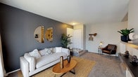 This single-floor Halsey Flats residence offers a thoughtfully designed layout with a contemporary aesthetic throughout. The living room immediately draws attention with its striking dark gray accent wall, adorned with a pair of round decorative mirrors. Plush carpeting, a jute area rug, and large windows dressed in soft linen curtains create a warm, inviting atmosphere. A dedicated entry area features a coat rack and a cozy reading nook with a leather accent chair.

The kitchen is equipped with white cabinetry, granite-look countertops, hardwood-style flooring, and black appliances including an electric range and built-in microwave. It opens directly into a casual dining area, providing a seamless flow for everyday living.

The primary bedroom is generously proportioned, featuring soft carpet, a ceiling fan, and a bold dark accent wall that frames the room with sophistication. Additional bedrooms are equally well-sized, each with plush carpet, ceiling fans, natural window light, and bifold closet doors offering ample storage.

Throughout the unit, neutral tones, modern fixtures, and consistent carpeting lend a cohesive and polished feel, making Halsey Flats a comfortable and functional place to call home.