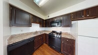 This single-floor residence offers a functional and comfortable layout with a seamless flow between living and kitchen areas. The open-concept design connects the carpeted living space to the kitchen via a partial dividing wall, which also features built-in shelving — ideal for display or storage.

The kitchen is a standout area, outfitted with rich, dark-stained wood cabinetry that extends to the upper walls, offering generous storage. Granite-style countertops provide a durable and attractive workspace, complemented by a full suite of black appliances including a gas range, built-in microwave, dishwasher, and a white refrigerator. Warm hardwood flooring and a bright window enhance the kitchen's inviting atmosphere.

The bedrooms are spacious and carpeted, each equipped with a ceiling fan for year-round comfort. One bedroom benefits from direct outdoor access through a sliding glass door, as well as a window that brings in natural light. Additional sleeping quarters are well-proportioned with clean white walls and neutral carpeting.

The bathroom is neatly appointed with tile flooring, a wood vanity topped with a granite-style countertop, a large wall mirror with overhead lighting, and a shower-tub combination. Ceiling fans are present throughout the living areas, contributing to a consistently comfortable environment across the entire residence.