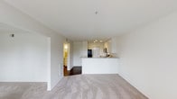 This single-floor space is designed with an open-concept layout that promotes a sense of spaciousness and ease of movement. The living area features plush wall-to-wall carpeting that adds warmth underfoot, while the kitchen and adjacent hallway transition to rich, dark hardwood flooring, creating a natural visual distinction between the two zones. The kitchen is finished in crisp white cabinetry that extends to the ceiling, offering ample storage. A peninsula counter with a light stone-toned surface serves as both a functional workspace and a casual dining or social area, seamlessly connecting the kitchen to the living space. Recessed lighting above the kitchen illuminates the work surfaces, complementing the natural brightness that fills the unit. Stainless steel appliances, including a built-in microwave and refrigerator, are neatly integrated into the cabinetry. The white walls and white ceilings throughout contribute to a clean, airy aesthetic that keeps the space feeling open and versatile. A hallway visible from the main living area leads to additional rooms, hinting at a well-organized floor plan. Overall, the unit presents a modern, neutral palette that offers a welcoming and functional environment.