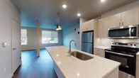 This single-floor unit offers a thoughtfully designed open-concept layout that seamlessly connects the kitchen and living areas, creating a bright and functional living space. The kitchen is a standout feature, showcasing light wood cabinetry that extends to the ceiling for generous storage, a suite of stainless steel appliances including a refrigerator, range, and microwave, and a sleek white quartz countertop island with an undermount sink and contemporary pendant lighting above. The backsplash is clean and minimal, complementing the modern aesthetic. Wood-look flooring runs consistently throughout the unit, adding warmth and a cohesive feel to every room. The open living area is bathed in natural light from large windows with roller shades, offering views of the surrounding neighborhood. The space also includes a large-format energy system unit, indicative of modern building infrastructure. The bathroom is cleanly appointed with a light wood vanity, white quartz countertop, and a glass-enclosed walk-in shower, all finished with matching wood-look flooring. Recessed lighting throughout ensures consistent, well-distributed illumination across the entire unit, contributing to the overall clean and contemporary ambiance.