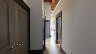 Located at 300 S Firestone St in Gastonia, NC, this thoughtfully converted loft unit masterfully blends industrial heritage with modern comfort. The open living area immediately commands attention with its soaring ceilings, warm wood-paneled ceiling, exposed ductwork, and original heavy timber beams that speak to the building's storied past. Polished concrete floors anchor the space, while oversized arched warehouse windows flood the area with abundant natural light and provide sweeping outdoor views.

The generously proportioned primary bedroom retains its industrial character through original wooden post columns and lofty ceilings, creating a dramatic yet intimate retreat. The second bedroom is equally impressive, featuring floor-to-ceiling steel-framed windows set against a striking exposed brick wall, with a ceiling fan adding a practical touch. Both bedrooms offer a warm, curated ambiance enhanced by the raw architectural elements.

A central hallway connects the bedrooms and three bathrooms with clean, efficient layouts. The bathrooms are finished with granite-topped vanities, warm wood cabinetry, and tub-shower combinations. Throughout the unit, the interplay of exposed brick, reclaimed timber, and crisp white walls creates a cohesive industrial-chic aesthetic that is both visually compelling and highly livable.