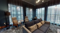 This sophisticated game room is designed for both relaxed lounging and engaging entertainment. The space is anchored by a striking wood-framed feature wall with a large mounted flat-screen television, set against dark, richly toned wood paneling that adds warmth and texture. A custom wooden media console beneath the screen provides ample storage and display space for games and accessories.

The room is furnished with a plush charcoal sofa accented with patterned throw pillows, complemented by a live-edge wood coffee table that adds a natural, artisanal touch. A pair of mid-century-style lounge chairs and a round bistro-height game table with seating near the windows create an inviting nook for tabletop games and conversation.

Floor-to-ceiling sheer dark curtains line an entire wall of windows, softening incoming natural light while maintaining a moody, intimate atmosphere. The deep teal wall color throughout enhances the dramatic ambiance, while a patterned area rug grounds the space underfoot. A standout architectural detail is the large sliding barn-style door with wood framing and textured glass panels, adding a refined industrial character. Track and pendant lighting provide layered illumination, completing this well-appointed, versatile entertainment retreat.