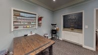 This single-floor space is thoughtfully designed as a multipurpose craft and gathering room, offering a warm and inviting atmosphere enhanced by soft blue-gray walls, white crown molding, and a textured ceiling with a ceiling fan and recessed lighting. The main area centers around a sturdy rustic wood worktable with attached stools, ideal for hands-on projects. A built-in white wall shelf displays an organized array of fabrics, ribbons, and craft supplies, keeping materials accessible and neatly arranged. A wall-mounted flat-screen television adds a multimedia element to the room. French doors fitted with white horizontal blinds allow natural light to filter in while providing privacy. An adjacent area features a chalkboard accent wall with white cabinetry and counter space, adding both a functional and creative touch to the room. A popcorn machine stationed nearby contributes a playful, social element. A cozy seating nook with striped armchairs and a small side table creates a comfortable lounge area, further enhanced by unique book-fold wall art that adds a literary, artsy character to the space. Patterned carpet flooring runs throughout, tying together the room's creative and welcoming ambiance. This well-appointed space is well-suited for crafting, socializing, and creative activities.