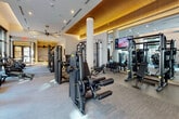 This contemporary wellness club is thoughtfully designed to support a wide range of fitness activities across a single, expansive floor. The space is defined by a clean, modern aesthetic, featuring a combination of light hardwood flooring in the entry and studio areas and durable carpet in the main equipment zones.

The main fitness area is generously outfitted with a comprehensive selection of professional-grade strength machines, free weights, a kettlebell rack, and multiple cable systems. Overhead, warm wood slat ceiling panels with integrated ambient lighting add an upscale architectural element, while large-blade ceiling fans ensure comfortable airflow throughout. Floor-to-ceiling grid-pane windows line the perimeter, flooding the space with natural light and offering views of an outdoor courtyard and the surrounding community.

A dedicated studio space features wall-to-wall mirrors, a ballet barre, hardwood floors, and an interactive touchscreen display for guided workouts. Plyometric boxes provide options for functional and high-intensity training. Adjacent to the studio, a bright transitional area houses a wall-mounted rack of medicine and stability balls, with direct access to the outdoor area via large glass doors.

The entry zone features warm pendant lighting, built-in cabinetry, and a welcoming lounge area, creating a polished, resort-style arrival experience that sets the tone for the entire facility.
