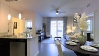 This single-floor residence features an open-concept layout that seamlessly connects the kitchen, dining, and living areas into one cohesive, light-filled space. The kitchen is outfitted with granite countertops and a breakfast bar that doubles as a casual dining surface, offering a natural transition into the dining area, where a round dark-toned table with upholstered chairs provides an elegant setting for meals. The living room is anchored by a ceiling fan with integrated lighting and features large sliding glass doors dressed with patterned drapes, allowing abundant natural light to pour in while offering direct access to an outdoor patio. The view through the glass doors reveals a green exterior space. Neutral-toned tile flooring runs throughout, tying the areas together and contributing to a clean, modern aesthetic. The overall ambiance is bright and welcoming, with a carefully curated décor palette of soft grays and whites that enhances the sense of openness. The layout is well-suited for both everyday living and entertaining.