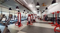This expansive single-level fitness center is designed with a modern industrial aesthetic, combining functionality and visual impact throughout its open layout. The space is anchored by a striking collection of bold red commercial-grade strength equipment, including cable machines, power racks, smith machines, and a comprehensive free weights area with barbells and adjustable benches. Floor-to-ceiling windows along one wall flood the cardio zone with natural light and offer views of the surrounding outdoor environment, where a row of treadmills and upright bikes are positioned to take full advantage of the scenery. Large pendant-style black dome lights hang from the exposed ceiling, complementing the industrial character of the space, while ceiling fans contribute to air circulation throughout. The flooring features a durable rubber tile pattern in a checkerboard arrangement, providing both safety and visual definition across different workout zones. Mirrored wall sections expand the sense of space and allow users to monitor their form during exercises. Near the entrance, a glass-partitioned lobby area and vending machines offer convenience for members. Concrete structural columns punctuate the floor plan, reinforcing the industrial design language that defines this well-equipped facility.