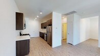 This single-floor space showcases a contemporary open-concept layout, with the kitchen serving as a central highlight. Dark espresso-toned cabinetry lines the kitchen walls, providing a striking contrast against the crisp white walls and light-toned hardwood-style flooring that extends throughout the unit. The kitchen is well-equipped with stainless steel appliances, including a refrigerator, dishwasher, and a built-in microwave, complemented by light-colored countertops that add brightness to the space.

Recessed lighting is installed throughout the ceiling, ensuring consistent, even illumination across the open living and kitchen areas. A structural column subtly defines the transition between the kitchen and the adjoining living space, maintaining an open feel while adding architectural interest. A ceiling-mounted HVAC vent ensures efficient climate control throughout.

A hallway visible beyond the main living area leads to additional rooms, with doors finished in white and accented by a warm yellow interior, hinting at a cheerful private space. The overall ambiance is clean, modern, and functional, with a neutral palette that offers a versatile backdrop for a variety of interior styles.