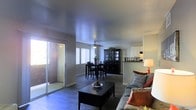 This single-floor residence offers an open and inviting layout that seamlessly connects the living and dining areas, creating a spacious and functional flow throughout the main living space. The living area is comfortably appointed, featuring wood-look flooring that extends throughout, providing a warm and cohesive aesthetic underfoot. A ceiling fan positioned above the dining area ensures year-round comfort, while large windows and a sliding glass door flood the space with natural light and provide convenient access to an outdoor patio or balcony area. The dining area accommodates a full dining set and is complemented by a striking dark wood china cabinet, adding both storage and a refined touch to the room. Just beyond the dining space, a glimpse into the kitchen reveals white cabinetry, hinting at a clean and bright cooking space. The neutral wall tones throughout create a calm, versatile backdrop that suits a variety of décor styles. The overall ambiance of the home is warm and welcoming, with an easy, open arrangement that makes the space feel both practical and comfortable for everyday living.