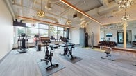 This fitness center combines contemporary design with a comprehensive selection of exercise equipment, creating an inviting and functional workout environment. The open-plan layout is anchored by an exposed ceiling featuring prominent orange accent beams, industrial ductwork, and a striking arrangement of decorative globe pendant lights that add a sophisticated touch to the space.

Large windows line the perimeter walls, flooding the interior with abundant natural light and offering views of the surrounding area. The flooring consists of patterned carpet tiles in shades of grey, teal, and yellow, defining distinct zones throughout the space. Floor-to-ceiling mirrors along one wall visually expand the room and provide practical utility for form-checking during workouts.

The facility is well-stocked with a diverse range of equipment, including treadmills, elliptical machines, indoor cycling bikes, and a cable strength-training machine. A dedicated free weights area features a full rack of dumbbells neatly arranged on tiered storage. A flat-screen television is mounted on the wall, and an adjustable workout bench provides additional versatility. Fitness center policies are posted on the walls, and the space is accessible through a glass-paneled door. The overall ambiance is bright, energetic, and modern, making it a well-appointed facility for a thorough workout.