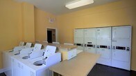 This shared laundry facility is a practical and efficiently organized space designed to serve multiple users with ease. The room is finished with warm yellow walls that create a bright and welcoming atmosphere, complemented by overhead fluorescent lighting that ensures excellent visibility throughout. A row of top-loading washing machines lines one side of the room, while a bank of stacked commercial dryer units occupies the opposite wall, providing substantial laundry capacity. Centrally positioned folding counters offer generous surface area for sorting and folding laundry, making the workflow intuitive and convenient. The layout is thoughtfully designed to allow users to move freely between machines and folding stations without congestion. A clearly marked exit sign and doorway provide easy access to adjacent areas, contributing to the room's safety and accessibility. The overall design prioritizes functionality, with durable, easy-to-clean surfaces throughout. This laundry room is a well-maintained, communal amenity that meets the everyday needs of its users in a clean and orderly environment.