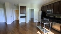 This single-floor space offers a practical and well-appointed layout designed for everyday comfort. The open-plan kitchen and living area is anchored by rich, dark hardwood-style flooring that runs throughout, creating a warm and cohesive aesthetic. The kitchen is equipped with dark espresso-toned cabinetry providing ample storage, a stainless steel refrigerator, microwave, and range, along with a stainless steel prep table that adds a functional, utilitarian touch. Dark countertops complement the cabinetry and contribute to a clean, modern look. A dedicated built-in cabinet nook with a dark countertop surface offers additional storage or workspace. The layout opens to a walk-in closet, adding generous storage capacity rarely found in similarly scaled spaces. A bathroom is also accessible from the main living area, featuring essential fixtures and a bright, clean finish. Natural light filters in through the space, highlighting the neutral green-toned walls that keep the interior feeling open and airy. The single-floor design ensures easy navigation throughout, making this a highly functional and efficiently organized space.