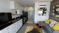 This single-floor unit showcases an open-concept layout that seamlessly integrates the kitchen and living area into one cohesive space. The kitchen is outfitted with white shaker-style cabinetry, dark granite countertops, and a full suite of black appliances, including a refrigerator, oven, microwave, and dishwasher. A stainless steel sink sits beneath upper cabinets that extend to the ceiling, maximizing storage capacity. The living area flows naturally from the kitchen and features a wall-mounted split air conditioning unit, providing efficient climate control. Bold yellow and white geometric-patterned area rugs anchor both zones, adding a vibrant contrast to the neutral grey flooring throughout. The living space is furnished with a grey sectional sofa, a warm wood-toned coffee table, and a framed abstract artwork that adds a contemporary artistic touch. A narrow entryway corridor with a patterned runner leads to the front door, offering a clear delineation between the unit's interior and entrance. The overall ambiance is modern and functional, with a cohesive color palette of white, grey, and yellow accents throughout.