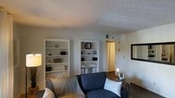This single-floor residence offers a well-organized and functional layout designed for comfortable everyday living. The living room serves as the central gathering space, featuring rich dark hardwood flooring that contrasts elegantly with the bright white walls and textured ceiling. Two sets of built-in shelving units are recessed into the wall, providing ample display and storage space while contributing a structured, built-in aesthetic to the room. The shelves are styled with decorative accents including bowls, a mirror, and small figurines, adding character to the space. A navy blue sofa anchors the seating area, complemented by a sleek side table. A framed wall mirror on the adjacent wall adds depth and visual interest to the room. A centrally placed ceiling vent ensures climate control throughout the space. A doorway leads from the living area into what appears to be an additional room, offering a glimpse of continued living space beyond. The overall ambiance is neutral and understated, with a clean, contemporary feel that makes the space feel organized, bright, and move-in ready.