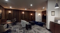 This stylish single-floor amenity lounge is thoughtfully designed to offer a refined yet comfortable social environment. The space is defined by its striking floor-to-ceiling vertical wood slat accent walls, which lend a warm, cinematic ambiance throughout. Recessed ceiling lighting is complemented by elegant wall sconces, casting a soft, inviting glow across the room.

The main lounge area is furnished with plush navy blue sofas, upholstered swivel armchairs, and dark accent tables, arranged to encourage both intimate conversation and communal viewing. A large flat-screen television is mounted against the wood slat feature wall, flanked by decorative artwork with a vintage-inspired typographic motif, adding a playful yet sophisticated character to the space.

A secondary seating cluster with a round dining table and chairs provides an area suited for casual meetings or social dining. The patterned carpet tiles throughout add visual texture and tie together the overall aesthetic.

Adjacent to the main lounge is a well-appointed kitchenette featuring dark wood cabinetry, stone countertops, a sink, and dramatic pendant lighting — ideal for entertaining. The overall design strikes a balance between a cozy screening room and an upscale social lounge, making it a versatile and welcoming communal space.
