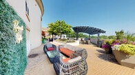 Located at 6205 Carnegie Blvd in Charlotte's SouthPark area, this rooftop terrace is a well-appointed outdoor amenity space offering a compelling blend of leisure, social gathering, and scenic views. The terrace is laid out across a curved, expansive footprint finished with herringbone-patterned brick pavers that lend a refined, cohesive aesthetic throughout.

The space is organized into several distinct zones. A central lounge area features stylish wicker-frame furniture with bold orange cushions, complemented by glass-top coffee tables and decorative lattice privacy screens that define gathering spots without obstructing sightlines. A standout feature is a green living wall panel adorned with a neon "Raise the Rooftop" sign, adding a vibrant, contemporary focal point to the space.

An outdoor pool with surrounding lounge chairs offers a resort-style retreat, while shaded dining areas with market umbrellas provide comfortable spots for al fresco meals. Mature ornamental trees and large planters with lush greenery are thoughtfully distributed to add warmth and a natural element to the rooftop environment.

The terrace benefits from abundant natural sunlight and open skies, and from the western edge, panoramic views of the Charlotte skyline stretch across the horizon, making it a particularly striking vantage point. The overall ambiance is upscale, inviting, and well-suited for both relaxed lounging and social gatherings.