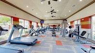 This fitness center at 14695 Briar Forest Dr offers a generously sized, single-floor workout space designed to accommodate a comprehensive range of exercise needs. The room features soaring vaulted ceilings equipped with multiple ceiling fans, recessed lighting, and crown molding, creating an open and airy atmosphere. Bold horizontal stripes of red, gold, and cream adorn the walls, giving the space a vibrant and energetic character.

Large windows line the perimeter, flooding the interior with abundant natural light while providing views of the surrounding landscaping. The floor is covered in a modern patterned carpet with orange and neutral tones, adding to the dynamic aesthetic.

The facility is comprehensively outfitted with a full suite of cardio equipment, including treadmills, ellipticals, and stationary bikes by brands such as Cybex. The strength training area includes a multi-station cable machine, weight benches, a dumbbell rack, and a selection of stability balls neatly stored on a dedicated rack. Wall-mounted televisions are positioned throughout for entertainment during workouts.

A water fountain is conveniently accessible, and a decorative herringbone-tiled accent wall adds a distinctive architectural touch to one corner of the room. The overall ambiance is energetic yet functional, making this a well-appointed fitness destination.