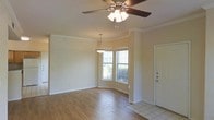 This single-story space showcases a thoughtfully designed open-plan living and dining area that combines comfort with classic architectural details. The living area is finished with warm hardwood-style flooring that extends throughout, contrasting with the tiled entryway near the front door. Crown molding runs along the perimeter of the ceiling, lending a refined, traditional character to the interior.

A ceiling fan with a built-in light fixture serves the living area, providing both airflow and ambient lighting. The dining area is defined by a elegant pendant light suspended from above, creating a natural gathering point. A curved bay window in the dining nook allows generous natural light to flood the space, offering views of the surrounding greenery outside.

The walls are finished in a neutral, warm beige tone that complements the flooring and trim, creating a cohesive and inviting atmosphere throughout. The front entry, delineated by tile flooring, transitions smoothly into the main living space, providing a practical and welcoming introduction to the home. Overall, the space feels open, bright, and move-in ready.