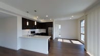 Located at 100 Citronica in Lemon Grove, this single-story space showcases a clean, modern aesthetic with an open-concept design that seamlessly connects the kitchen and living areas. The kitchen is a standout feature, equipped with dark espresso cabinetry that contrasts sharply against bright white countertops, creating a bold and stylish look. Stainless steel appliances, including a refrigerator, range, and microwave, complement the contemporary design. A peninsula with a white countertop provides additional workspace and casual seating, making it ideal for both cooking and entertaining. Pendant lighting hangs above the peninsula, adding focused illumination and a design accent. Recessed lighting throughout the ceiling ensures the space remains well-lit. Rich, dark hardwood-style flooring runs throughout, grounding the space with warmth and continuity. The open living area beyond the kitchen is bright and airy, benefiting from natural light that streams in through a window visible in the adjacent room. Crisp white walls enhance the sense of openness, while the overall layout offers a functional and inviting environment with a distinctly modern character.
