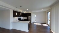 Located at 100 Citronica in Lemon Grove, this single-story space showcases a clean, modern aesthetic with an open-concept design that seamlessly connects the kitchen and living areas. The kitchen is a standout feature, equipped with dark espresso cabinetry that contrasts sharply against bright white countertops, creating a bold and stylish look. Stainless steel appliances, including a refrigerator, range, and microwave, complement the contemporary design. A peninsula with a white countertop provides additional workspace and casual seating, making it ideal for both cooking and entertaining. Pendant lighting hangs above the peninsula, adding focused illumination and a design accent. Recessed lighting throughout the ceiling ensures the space remains well-lit. Rich, dark hardwood-style flooring runs throughout, grounding the space with warmth and continuity. The open living area beyond the kitchen is bright and airy, benefiting from natural light that streams in through a window visible in the adjacent room. Crisp white walls enhance the sense of openness, while the overall layout offers a functional and inviting environment with a distinctly modern character.