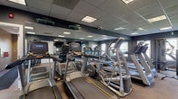 Located at 842 Cobblestone Cir in Painesville, Ohio, this fitness center is a well-equipped, single-floor facility designed to support a full range of workout routines. The space is finished with a drop tile ceiling outfitted with recessed fluorescent lighting panels that ensure consistent, bright illumination throughout. Deep blue-gray walls give the interior a focused, energetic atmosphere, while large wall-mounted mirrors in the main cardio area visually expand the space and allow users to monitor their form during exercise.

The cardio zone is generously stocked with multiple Matrix and Titan treadmills, elliptical trainers, and stair climbers, all arranged to maximize usability and flow. Natural light filters in through several windows along the perimeter walls, offering views of the surrounding outdoor areas. A dedicated strength training section includes a cable/pulley machine, adjustable benches, a dumbbell rack, and a stationary recumbent bike, providing options for both resistance and low-impact training.

The flooring transitions between durable carpet tile in the main exercise areas and hard tile near the entry, making for easy maintenance. Wall-mounted televisions are positioned throughout for entertainment during workouts. Large glass windows and a doorway connect the fitness center to an adjacent lounge or clubhouse area, enhancing the sense of openness and community within the facility.