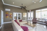This thoughtfully designed single-floor space serves as an inviting communal retreat, combining functional amenities with stylish interior finishes throughout. The main lounge area makes a strong first impression with its vaulted ceilings, warm hardwood flooring, and rich, dark-toned walls that create a sophisticated ambiance. A ceiling fan anchors the space overhead, while large floor-to-ceiling sliding glass doors flood the interior with abundant natural light and offer direct views of an outdoor pool and patio area, seamlessly connecting indoor and outdoor living. Comfortable patterned accent chairs and a neutral area rug define the seating arrangement, complemented by contemporary artwork on the walls. Adjacent to the lounge, a dedicated dining area with dark wood furniture provides an additional gathering space beneath a bright window. A secondary lounge area features a cream leather sectional sofa with bold accent pillows, framed by tall ceilings with crown molding and elegant French doors that open into the kitchenette. The kitchenette is well-equipped with light maple cabinetry, stainless steel appliances including a full-size refrigerator, a mosaic tile backsplash, and dark countertops — all set atop continuous hardwood flooring that ties the entire space together cohesively.
