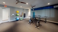 Located in Riverview, FL, this single-floor fitness facility at 9511 Wes Kearney Way offers a thoughtfully designed workout space divided into distinct functional zones. The interior features neutral gray walls, dark carpeted flooring, and recessed lighting throughout, creating a clean and focused environment conducive to exercise.

The main workout area houses a row of stationary cycling bikes positioned beneath ceiling fans that promote airflow during intense sessions. A wall-mounted flat-screen television is positioned for visibility across the room, adding an entertainment element to cardio workouts. Interior windows connect this space to an adjoining room equipped with treadmills, allowing natural sightlines between areas.

A dedicated functional training zone features an organized equipment rack stocked with resistance bands, medicine balls, stability balls, and foam rollers. A wall-mounted exercise mat provides additional versatility for stretching and floor-based routines. The space also includes a cable machine for strength training. Exposed blue ductwork runs along the upper portion of the walls, adding an industrial accent to the otherwise polished interior. The overall layout is open and uncluttered, maximizing usable floor space for a variety of fitness activities.
