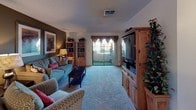 This single-floor space centers around a well-appointed living room that balances comfort and functionality. The room is anchored by a large wooden entertainment center housing a flat-screen television, complemented by matching pine-toned bookshelves that provide both storage and display space. Plush wall-to-wall carpeting in a neutral tone runs throughout, contributing to the room's warm and inviting atmosphere.

Seating is arranged thoughtfully, with a blue sofa accented by colorful throw pillows, a patterned armchair, and a dark wood coffee table at the center. The accent wall, painted in a deep earthy brown, adds depth and contrast to the otherwise light interior. Framed artwork adorns the walls, adding a personalized touch.

A sliding glass door fitted with draped curtains leads to a private outdoor balcony, allowing natural light to filter into the space and offering a pleasant outdoor view. The textured ceiling features a centrally located HVAC vent, ensuring climate control throughout. Decorative plants add a lively, organic element to the overall aesthetic.