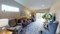 This single-floor residence offers a well-organized layout with distinct living spaces connected by a cohesive, warm design aesthetic. The living room features vaulted ceilings, a patterned area rug over carpet, and a sliding glass door that opens to an outdoor patio with views of a green landscape. Framed equestrian artwork and mixed seating, including leather and upholstered chairs, contribute to a comfortable, country-inspired atmosphere.

The kitchen is generously sized with rich medium-toned wood cabinetry, granite-look countertops, a mosaic tile backsplash, and durable wood-look flooring. White appliances and ample counter space make this a highly functional cooking area. Adjacent to the kitchen, the dining area features hardwood-style flooring and opens to a taller ceiling section, giving the combined space an airy feel.

The primary bedroom is spacious with plush carpeting, a window overlooking the outdoors, and room for substantial furniture. A secondary bedroom offers carpeted flooring and built-in storage. Both bathrooms are finished with granite-topped dark wood vanities and tiled flooring, providing a consistent, polished look throughout the home.