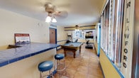 This single-floor multipurpose space seamlessly combines recreation and fitness in a colorful, inviting environment. The room is defined by warm yellow walls accented with a bold blue horizontal stripe, creating a lively and energetic atmosphere throughout. Terracotta tile flooring spans the entire space, adding a warm, earthy tone that complements the vibrant color scheme.

At the heart of the room sits a classic wooden pool table, serving as the centerpiece for social and recreational activity. Adjacent to it, a tiled bar counter in deep blue is paired with matching blue barstools, offering a casual spot to relax and socialize. The fitness area is well-equipped with a treadmill, elliptical, and additional exercise machines, making it easy to maintain an active lifestyle without leaving the space.

Large windows along one wall flood the room with natural light, enhancing the open and airy feel. Ceiling fans are positioned throughout to ensure comfortable airflow. Decorative murals or artwork on the far wall add a distinctive personal touch and visual interest to the space. A private door provides additional access or storage. Overall, this space offers a well-rounded environment designed for both fitness and fun.
