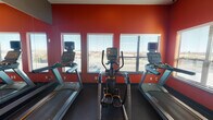 Located at 319–347 Nichols St in Fort Worth, Texas, this single-level fitness center is designed to support a comprehensive range of workout routines. The space is divided into distinct functional zones, each thoughtfully appointed with modern equipment and a cohesive, energizing aesthetic defined by bold red and orange accent walls paired with neutral gray tones and dark flooring.

The cardio area houses multiple Matrix treadmills and an elliptical trainer, all positioned to face a row of windows fitted with horizontal blinds. These windows allow ample natural light to fill the space while offering views of the surrounding area, creating an open and airy atmosphere during workouts. A ceiling fan and recessed lighting provide additional comfort and visibility.

Floor-to-ceiling mirrors are strategically placed throughout the facility, visually expanding the space and allowing users to monitor their form. A separate functional training zone features spinning bikes, stability balls, foam rollers, resistance equipment, and neatly organized free weights displayed on wall-mounted racks. Colorful artwork adorns the walls, adding a dynamic visual element to the space.

Glass-paneled doors connect the different workout zones, maintaining an open, connected feel while allowing clear sightlines throughout. The overall design balances practicality with an energetic ambiance well-suited for focused, motivating exercise sessions.