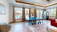 Located in South Ogden, Utah, this single-floor community clubhouse offers a well-appointed and versatile gathering space designed for both relaxation and recreation. The open-plan layout is anchored by light wood-look flooring throughout, creating a bright and cohesive aesthetic.

The living and lounge area is a standout feature, boasting a tray ceiling accented with exposed wood beams and ambient recessed lighting. A built-in shelving unit with integrated display niches frames a wall-mounted flat-screen TV and a sleek linear fireplace below, providing a stylish focal point. The space comfortably accommodates both lounge seating and recreation, with room for a pool table and a ping pong table. Large windows and glass doors flood the area with natural light and offer direct views of the outdoor pool and surrounding community.

The kitchen is well-equipped with rich dark wood cabinetry, white quartz countertops, a geometric tile backsplash, and a central island with bar seating illuminated by a trio of pendant lights. Ample counter space, a stainless steel refrigerator, and a double sink make this an ideal space for entertaining. Natural light pours in through large windows, enhancing the kitchen's welcoming atmosphere. The overall design blends modern finishes with warm, natural elements for a functional and inviting community space.