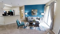 This single-floor residence makes a bold design statement through a thoughtfully curated interior that balances color, comfort, and functionality. The living area serves as the centerpiece of the home, anchored by a striking teal accent wall that adds depth and personality to the space. Soft carpeted flooring runs throughout, contributing to a warm and inviting ambiance. Large sliding glass doors allow natural light to pour in, brightening the room and offering a connection to the outdoors. The open layout flows naturally from the living area toward the kitchen, which is visible through a pass-through counter bar, creating a sense of connectivity ideal for entertaining. The kitchen features cabinetry in a warm wood tone and pendant lighting that adds a subtle decorative touch. The living room is well-appointed with a neutral-toned sofa, a teal accent chair, mirrored side tables, and a geometric area rug in coordinating tones of teal, grey, and white, all of which complement the bold accent wall. A piece of abstract artwork displayed prominently above the sofa ties the color palette together cohesively. The overall ambiance of the space is modern and curated, with a careful attention to color coordination and design detail throughout.
