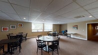 Located at Campus Edge in Carbondale, IL, this single-floor community space is thoughtfully designed to serve a variety of social and functional needs. The main gathering area features multiple round and rectangular tables paired with cushioned chairs, making it well-suited for dining, studying, or group activities. Warm beige walls, carpeted flooring, and a drop tile ceiling with recessed lighting create a comfortable, welcoming atmosphere throughout.

Large windows with vertical blinds line the walls, allowing natural light to fill the space while offering views of the surrounding greenery. Colorful framed artwork adds a lively touch to the walls, enhancing the communal feel of the area.

A dedicated lounge area is furnished with rich leather sofas, a large wall-mounted flat-screen television, and a foosball coffee table, providing a relaxed setting for recreation and socializing. An adjacent quieter room offers a plush armchair and a glass side table beside oversized windows, ideal for reading or unwinding.

A carpeted corridor connects the various areas of the space, with ceiling-mounted HVAC units ensuring year-round comfort. Additional utility areas, including what appears to be a small workstation with a computer, round out the practical offerings of this well-equipped community facility.