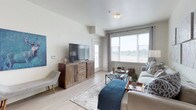 This well-appointed single-floor residence offers a thoughtfully designed layout with modern finishes and a clean, contemporary aesthetic throughout. The open-concept kitchen is a standout feature, showcasing sleek white upper cabinetry, stainless steel appliances including a refrigerator, microwave, and range, light wood-look flooring, and a large island with a white countertop and bar seating — ideal for casual dining or entertaining. Pendant lighting above the island adds a stylish finishing touch.

The living room is bright and welcoming, benefiting from generous natural light streaming through large windows with outdoor views. Light hardwood-style flooring flows seamlessly through the main living areas, lending a sense of openness and continuity.

Two well-proportioned bedrooms feature plush carpeting, large windows with soft drapery, and a calm, neutral palette that promotes comfort and relaxation. Each bathroom is finished with tile flooring, a combined tub and shower with white subway-style tile surround, and a vanity with a dark wood-grain cabinet and white countertop.

A dedicated in-unit laundry area houses a full-size top-loading washer and dryer with overhead shelving, adding everyday convenience. The overall design balances functionality with modern style, creating a cohesive and comfortable living environment.