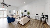 Located at 108 Lords Court in Virginia Beach, VA, this single-floor community clubroom is a well-appointed gathering space designed for both relaxation and recreation. The open-concept layout is anchored by a crisp, all-white interior that exudes a clean and contemporary aesthetic. Elegant paneled wainscoting lines the walls throughout, adding a refined architectural detail that elevates the room's overall character.

At the heart of the lounge area sits a classic white fireplace framed by a traditional mantel, with a wall-mounted television positioned above — creating an inviting focal point. Rich, hardwood-style plank flooring runs the length of the space, providing warmth and contrast against the bright white walls. Generous seating arrangements, including sectional sofas and accent chairs, establish a comfortable social atmosphere.

The space also features a full-size pool table, offering a dedicated recreational zone ideal for community gatherings. A high ceiling with a ceiling fan ensures comfortable airflow throughout. Multiple windows along the perimeter flood the interior with natural light, while French doors at the entry provide a welcoming, open feel with views to the exterior. A bar-height table with seating and a console table near the entrance complete the versatile, thoughtfully arranged layout.