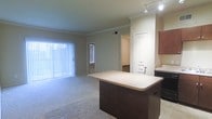 This single-floor residence is designed with an open-concept layout that seamlessly connects the kitchen and living areas, creating a sense of spaciousness and ease of movement throughout. The kitchen features a central island with a light-toned countertop and dark wood cabinetry, offering both prep space and a natural dividing point between the cooking and living zones. Track lighting above the kitchen provides focused illumination, complementing the overall warm, neutral color palette of the walls and finishes. The living area is finished with plush carpeting that extends throughout, lending a comfortable and cohesive feel to the space. Large vertical-blind-covered sliding glass doors dominate one wall of the living area, allowing generous natural light to filter in and providing access to an outdoor area. Crown molding along the ceiling adds a refined architectural detail. A secondary windowed door is also visible, contributing additional light. Beyond the main living space, a hallway leads to additional rooms, hinting at a practical and well-organized floor plan. The overall ambiance is clean, neutral, and functional, with a layout well-suited for everyday living.