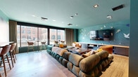 Located at 301 Winchester Ave in New Haven, CT, this single-floor lounge space offers a thoughtfully designed and visually striking communal environment. The room is defined by its signature teal-painted walls and matching ceiling, which lend a cohesive, immersive atmosphere throughout. A large-scale artistic wall mural featuring birds and abstract shapes in white, yellow, and purple serves as a dramatic focal point, adding a creative, gallery-like quality to the space.

At the heart of the room sits an expansive modular sectional sofa in deep teal and warm brown velvet, arranged around a solid wood coffee table — ideal for gathering and relaxation. Complementing the seating arrangement are cognac leather accent chairs that introduce warm contrast to the cool color palette. Recessed lighting in the ceiling provides even, ambient illumination throughout the space.

A wall-mounted flat-screen television is paired with a built-in media console, creating a dedicated entertainment zone. A bar-height counter along the back wall, lined with cognac leather bar stools, offers additional seating and a more casual dining or social setting. Floor-to-ceiling windows along one wall flood the space with natural light and frame views of the surrounding New Haven streetscape, connecting the interior to its vibrant urban context.