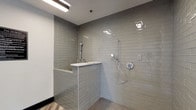 This single-floor space is centered around a thoughtfully designed, accessibility-focused shower room that combines clean aesthetics with practical functionality. The walls are clad floor-to-ceiling in glossy grey subway tiles, lending a bright, contemporary look that reflects light efficiently throughout the space. Overhead, a linear fluorescent fixture provides consistent, even illumination, complemented by a wall-mounted utility light that ensures the room is well-lit at all times.

The open, barrier-free shower area is designed with accessibility in mind, featuring a built-in tiled bench topped with a crisp white surface — ideal for seated use or convenient storage. Two handheld showerheads are mounted at varying heights on adjustable slide bars, offering flexible use for a wide range of users. The chrome fixtures add a polished, modern touch against the cool grey tile backdrop.

Underfoot, a classic penny tile floor in a neutral tone provides both visual texture and slip resistance, enhancing safety without compromising style. The transition to wood-look flooring at the room's threshold adds warmth and clearly delineates the wet zone. The overall ambiance is clean, functional, and modern — a well-appointed space designed to serve diverse needs with understated elegance.