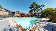 This inviting outdoor amenity space centers around a generously sized, freeform swimming pool with crystal-clear blue water, framed by a classic brick and wood architectural aesthetic. The pool area is enclosed by a wrought-iron security fence and flanked by mature trees, including tall pines and a palm, lending the space a resort-like atmosphere under open blue skies.

Adjacent to the pool is a covered pergola structure with a decorative lattice roof that casts striking geometric shadow patterns across the deck below. Ceiling fans provide added comfort in the shaded lounge area, which is furnished with dark wicker seating accented by teal throw pillows, arranged around a central coffee table — ideal for relaxing in the shade.

The wood-planked deck extends from the pergola toward the poolside, offering a seamless transition between shaded and sun-drenched areas. Multiple umbrella-shaded patio tables and chairs are positioned poolside for additional outdoor dining and lounging. A life ring mounted near the fence underscores the safety-conscious design.

A brick-clad leasing or amenity office with large multi-pane windows and a glass entry door sits directly adjacent to the pool deck, offering convenient access. The overall space is well-maintained, open, and thoughtfully designed for both relaxation and community enjoyment.