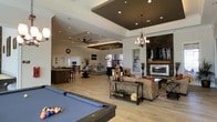 This expansive, single-floor open-concept space is designed to impress and accommodate. The layout flows seamlessly from one zone to the next, creating a versatile environment ideal for both socializing and relaxation. A full-size billiards table anchors the foreground, setting the tone for the recreational focus of the space.

The living area features plush seating arranged around an eye-catching stone fireplace with a modern linear insert, serving as a striking focal point along the far wall. Overhead, dramatic tray ceilings — some finished in a contrasting dark tone — add architectural depth and visual interest, complemented by a mix of pendant chandeliers and ceiling fans that provide both ambient and functional lighting.

Light wood flooring runs throughout, lending warmth and cohesion to the open floor plan. Toward the rear of the space, a wet bar or kitchenette with bar seating offers a convenient service area, while additional lounge furniture creates a secondary gathering zone bathed in natural light from large windows.

The overall ambiance is sophisticated yet inviting, blending industrial-chic accents — including metal-framed furniture and exposed stone — with comfortable, residential-style furnishings. This space is thoughtfully designed to serve as a premier social and recreational hub.