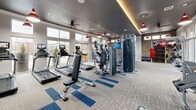 Located in Charlotte, NC, this fitness center offers a thoughtfully designed, single-floor workout facility that provides a comprehensive exercise experience. The space features a striking dark ceiling with warm ambient cove lighting and bold red pendant lights that contrast against light gray walls and an exposed brick accent wall, creating an energetic yet modern atmosphere.

The layout is open and spacious, accommodating a full range of fitness equipment across distinct zones. The cardio area includes treadmills, elliptical trainers, stair climbers, and stationary bikes, all positioned near large windows that flood the space with natural light and offer views of the surrounding outdoor area. A row of flat-screen TVs ensures entertainment during workouts.

The strength training section features a cable machine, a multi-functional rack with weight plates, and a selection of benches and free weights. A dedicated functional training zone in the rear houses a punching bag, medicine balls, kettlebells, battle ropes, TRX suspension trainers, and a RAX multi-use rig—ideal for high-intensity interval and cross-training workouts.

The flooring throughout consists of durable carpet tile with blue accent stripes, adding a dynamic visual element. Ceiling fans provide air circulation, while glass doors open to the outdoors. The overall design balances aesthetics with practicality, making this a versatile, full-featured fitness facility.