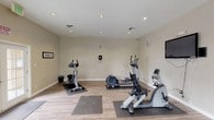 This fitness center is designed to support a well-rounded workout routine, offering a thoughtfully arranged, multi-room layout spread across a single floor. The space features light wood-look flooring throughout, recessed lighting, and neutral-toned walls accented by a distinctive teal feature wall that separates the two connected workout areas.

The primary room houses an assortment of cardio equipment, including multiple elliptical trainers and treadmills by SportsArt, each placed on protective rubber mats. A wall-mounted flat-screen television is positioned for convenient viewing during workouts. The secondary room provides additional cardio options, including a stationary bike and rowing machine, alongside a multi-function cable strength training station with an attached bench, catering to resistance and strength training needs.

Natural light filters in through several windows, offering views of an outdoor patio area complete with seating and umbrellas, creating an airy and motivating atmosphere. French doors provide access to the exterior, further enhancing the connection to the outdoors. The facility is equipped with a sanitization station, video surveillance for security, and framed instructional or motivational artwork on the walls. The overall ambiance is clean, functional, and inviting, making it a practical space for daily fitness activity.